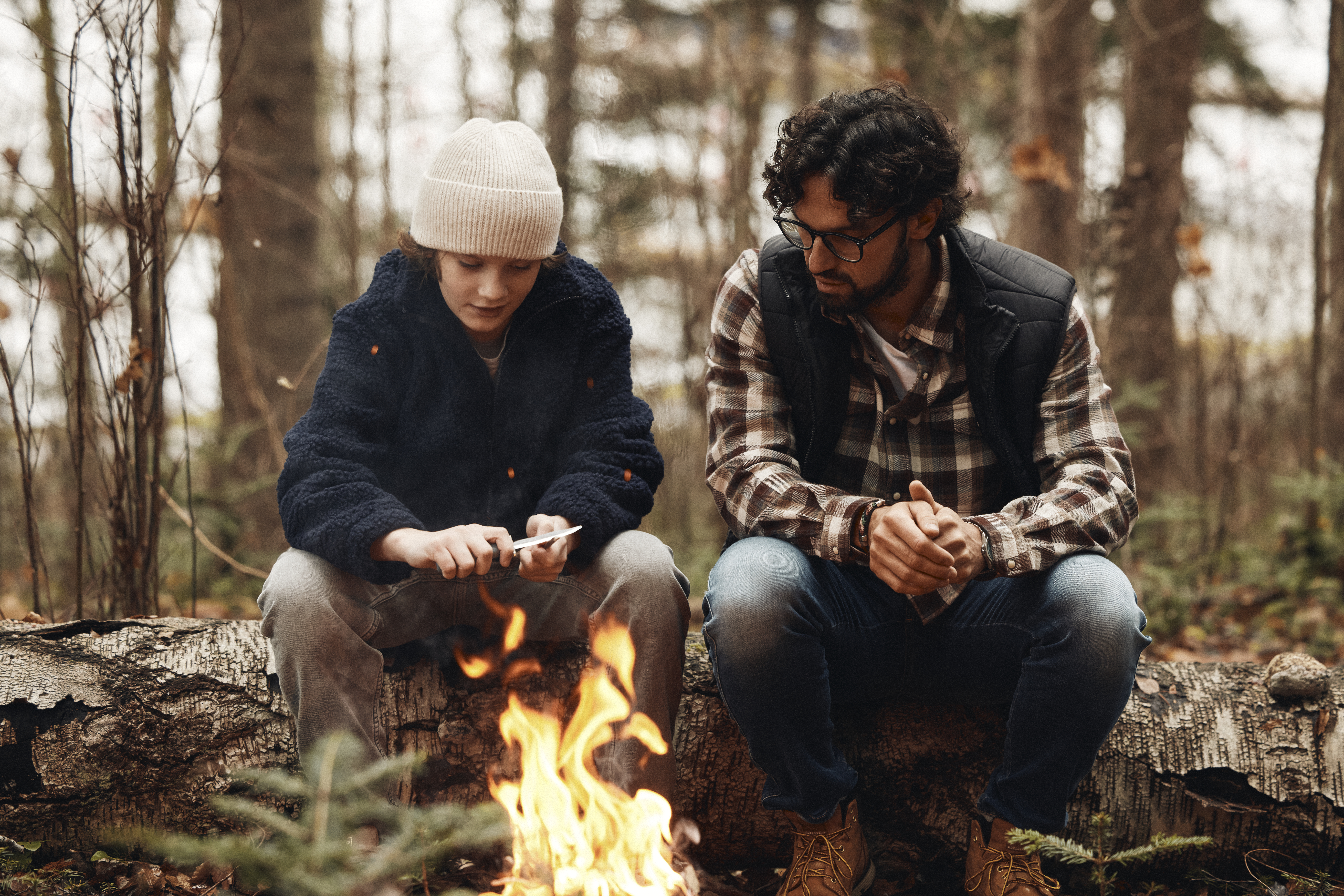 två personer som sitter på en trästock framför en brasa. en person täljer på en träbit med en kniv.